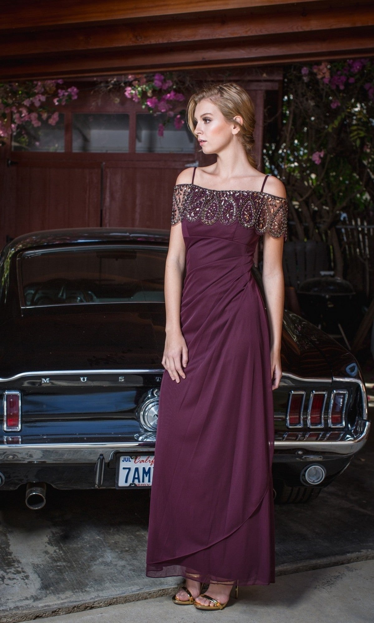 A woman in a burgundy Off-Shoulder Draped Long Formal Dress: Eureka 3937 poses by a classic black Ford Mustang in a garage adorned with flowers and greenery, dressed for a black-tie event.